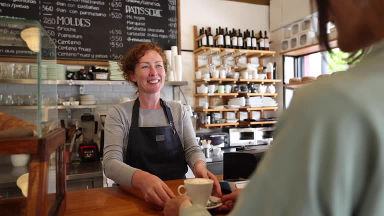 Happy barista working at a cafe serving a cup of coffee to businesswoman taking a break from work
