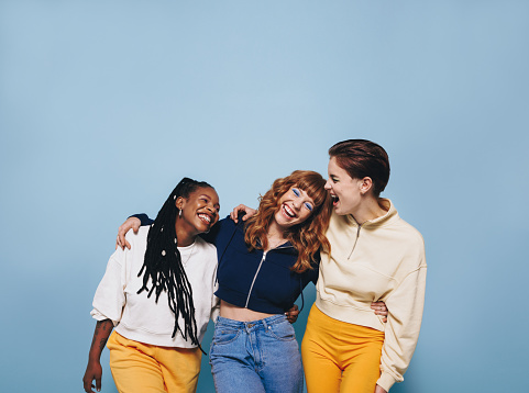 Multicultural female friends laughing and having fun while embracing each other. Group of cheerful young women enjoying themselves while standing against a blue background.