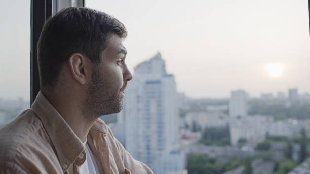 istock Sad middle-aged man standing by window looking at urban view, upset with a problem 1447417209