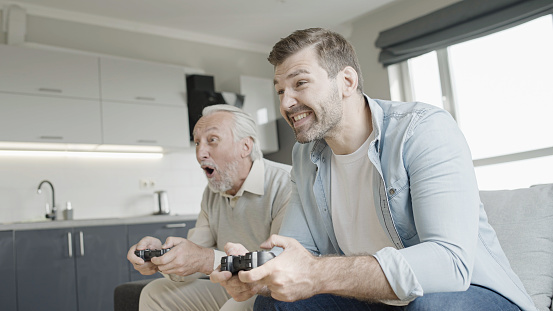Cheerful senior father and adult son playing video game at  home, having fun together