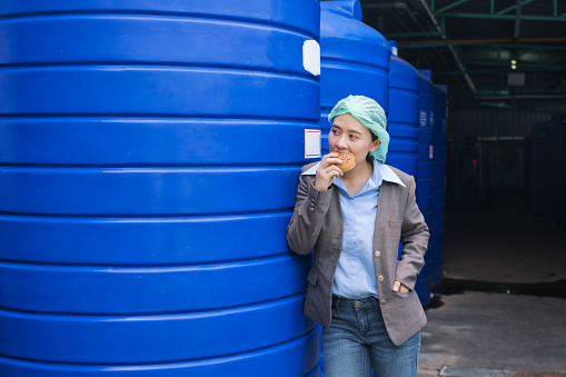 Young asian supervisor woman standing eating bread with haste during lunch break in water tank warehouse in processing factory