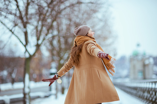 Seen from behind woman in brown hat and scarf in camel coat with gloves and raised arms rejoicing outside in the city in winter.