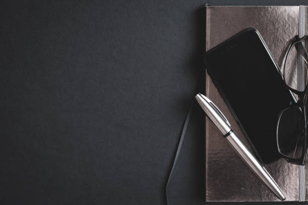 Shiny silver journal and silver pen with reading glasses and cell phone device as a border on a black background with copy space