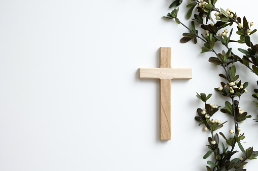 Small wood christian cross on a white background with a border of branches with green leaves and white berries shot from above with copy space
