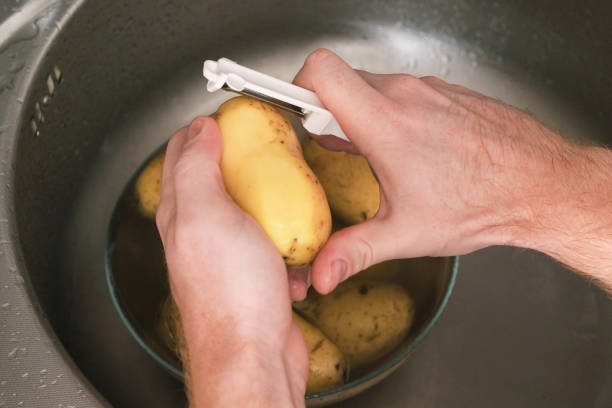 男が流しの野菜の皮むき器でジャガイモの皮をむきます。調理用ジャガイモの準備。農薬から皮をむきます。男性は家族のために家で夕食や昼食を作ります。 - root vegetable raw potato human skin root ストックフォトと画像