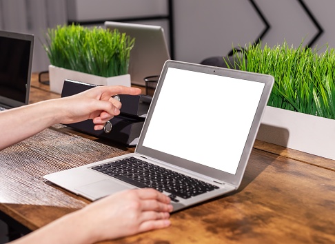 Finger pointing at blank white screen mockup of laptop computer in office interior. High quality photo
