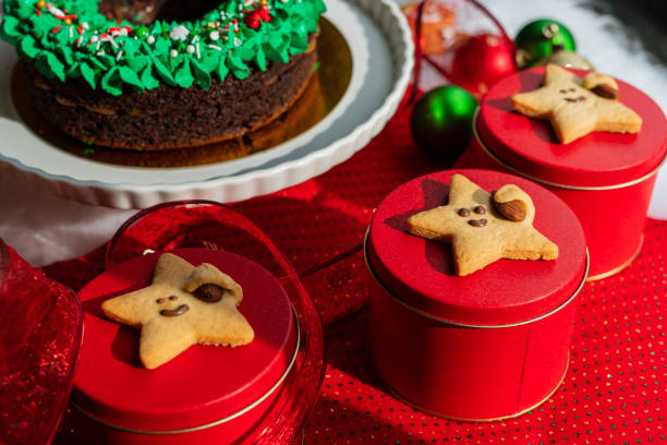 Plate of frosted Christmas cookies on checkered cloth, surrounded by red and green ornaments, gift boxes, and blurred background with fireplace and tree, cozy indoor holiday celebration setup.