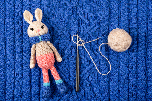 Knitted children's toy hare or rabbit with a ball of thread and a crochet hook on a knitted blue cloth. The symbol of 2023.