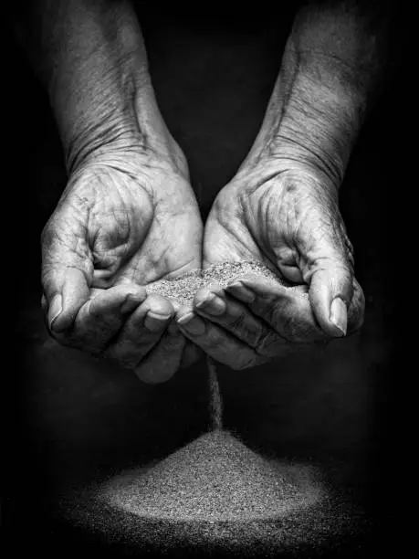 Photo of Sand between veteran hands like an hourglass. The concept of passing time. Hands weathered by time dropping sand like clockwork. Old hands synonymous with that Time is running out