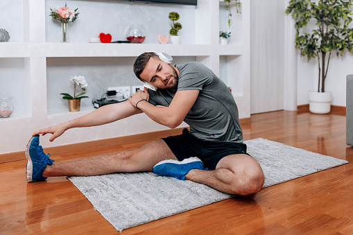 A young Caucasian man wearing headphones is sitting on an exercise mat and stretching.
