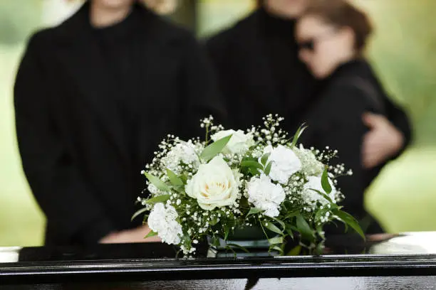 Close up of flower bouquet on coffin with people wearing black in background, outdoor funeral ceremony, copy space