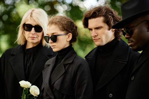 Group of people dressed in black standing at outdoor funeral ceremony with flowers