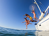 Group of friends diving in the water during a sailboat excursion, young people jumping inside ocean in summer vacation from a sail, having fun, luxury vacation lifestyle