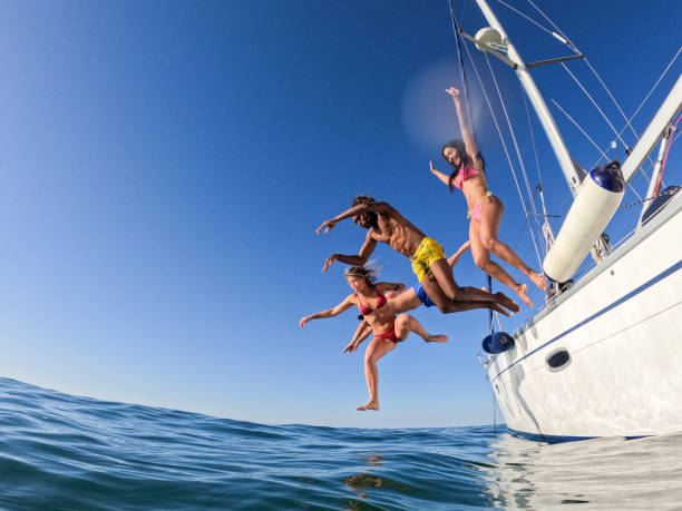 group of friends diving in the water during a sailboat excursion, young people jumping inside ocean in summer vacation from a sail, having fun, luxury vacation lifestyle - beach nautical vessel party clothing imagens e fotografias de stock