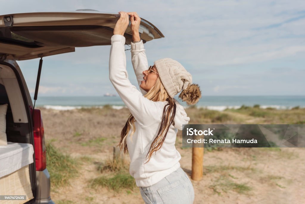 a woman with white winter bobble hat opening or closing the trunk door of her parked camper van close to the beach. Van life concept Closing Stock Photo