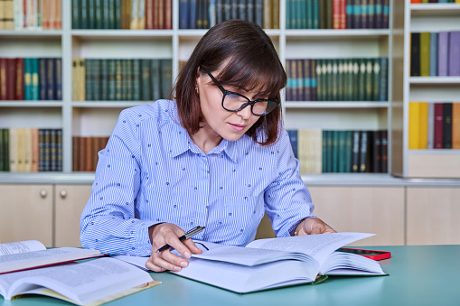 Middle-aged female teacher working in library, writing in papers at desk. Education, knowledge, school, college, university, technology, teaching concept