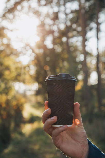 A black paper cup with coffee in a man's hand close-up on the background of the setting sun among the leaves of the trees stock photo