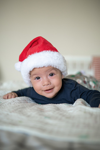 Adorable 6 months old newborn baby boy lying down on bad wearing Santa Claus hat and festive winter pajama set for New Year and Christmas holiday celebration