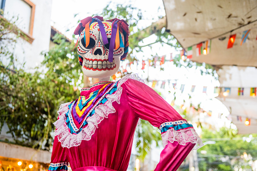 In Playa del Carmen, Mexico just after Day of the Dead a large skeloton statue with a sugar skull face and traditional clothing remains on display along 5th Avenue surrounded with palm trees.