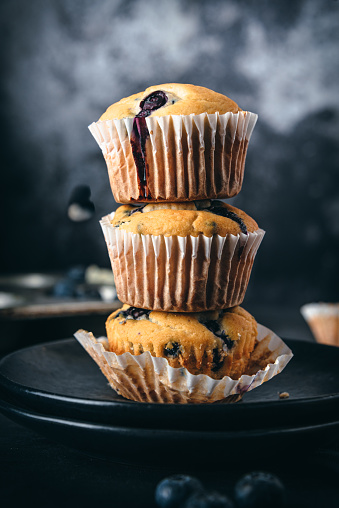 Stack of homemade blueberry muffins in paper cake cases surrounded by fresh blueberries, black background.