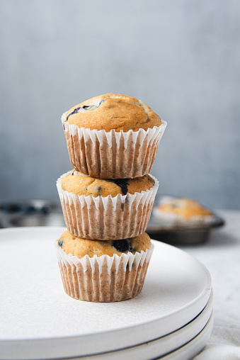 Stack of homemade blueberry muffins in paper cake cases surrounded by fresh blueberries, gray background.