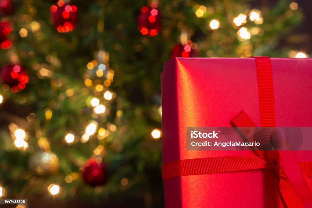 Gift and Christmas tree. Goiânia, Goias, Brazil – December 07, 2022: A gift box, red, with a ribbon bow, with a Christmas tree in the background. Backgrounds Stock Photo