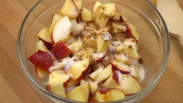 Sugar and cinnamon powder are sprinkled on sliced apples in a glass bowl