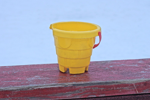 one yellow plastic bucket toy stands on a red wooden table outside on a white background
