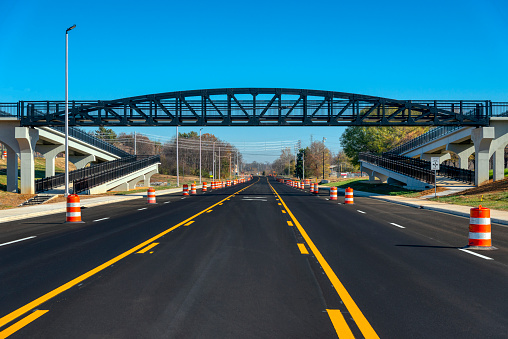 Newly-painted stripes and orange traffic barrels are seen on a freshly-blacktopped road under a pedestrian walkway bridge.