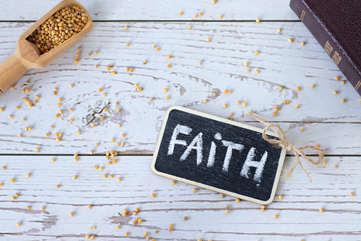 Faith handwritten text word with chalk, spoon with mustard seeds, and closed holy Bible book on wooden background. Top table view.