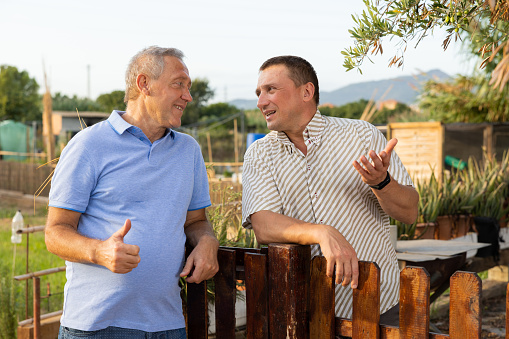 Two male amateur gardeners friendly talking outdoors next to wooden fence of country estate on summer day