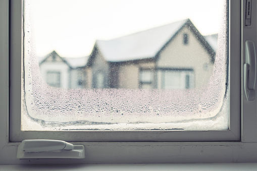 A large window is frozen over on a frigid winter morning in Canada. The temperature outside is -30 degrees Celsius and the condensation has frozen over the glass panels. There is snow outside the window and the sun is slowly rising.