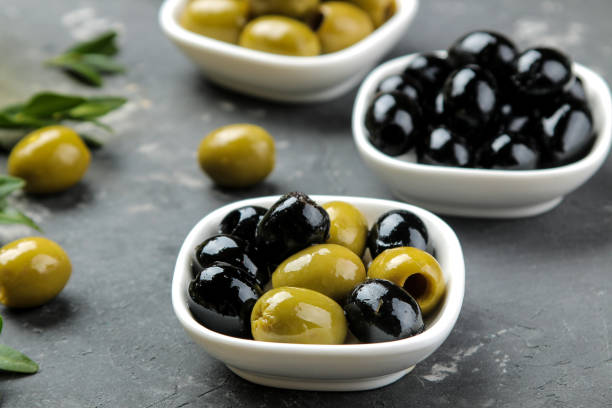 istock Green and black olives in a white ceramic bowl with leaves on a dark graphite background. close-up 1447524882