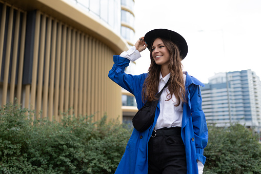 stylish young woman in an autumn blue coat and black hat with a smile on her face is walking near the mall.