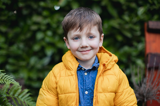 A cute little boy about 4-5 years old wearing a blue shirt with a yellow jacket. The photo is composed like a head shot showing mostly shoulders and head. Shot against a soft green natural plant background.