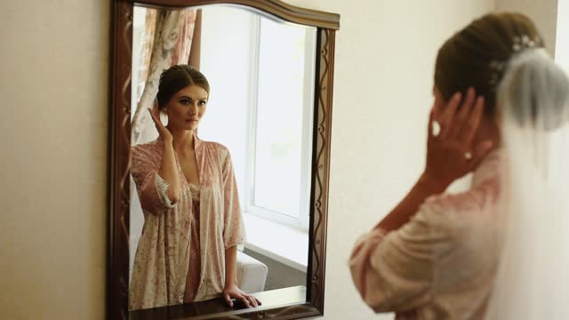 girl with veil is standing in pink nightgown and satin robe looking mirror