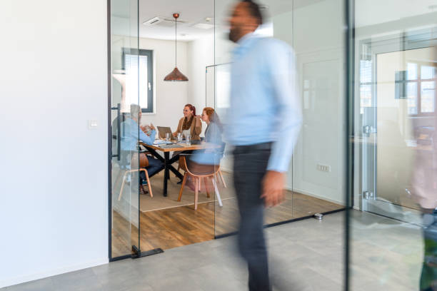 Diverse Group of Employees Moving Around Open Space Office Front action has people walking around open space office. The background includes a diverse group of employees at a business meeting. walking into glass door stock pictures, royalty-free photos & images