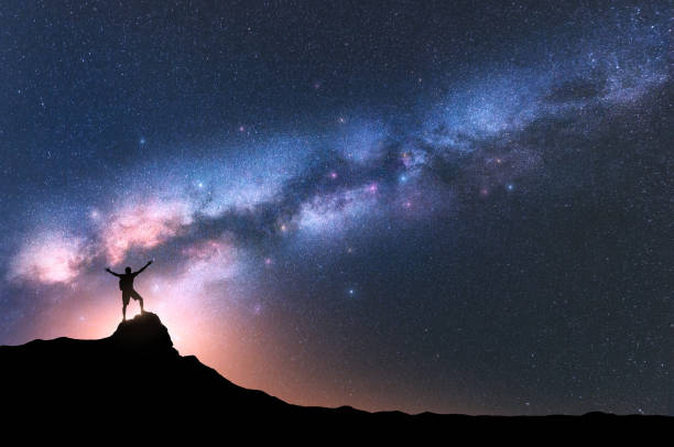 Milky Way and happy man with backpack on the mountain peak at night. Silhouette of guy with raised up arm on the hill, sky with stars, yellow light in Nepal. Galaxy. Space landscape with milky way stock photo