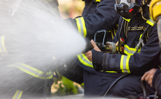 Firefighters in a fire protection suit wearing firefighter helmet with breathing device and holding fire hose is extinguishing a burning house fire that is putting off excessive heat and smoke.  Fire could have been caused by accident or arson.  Second firefighter in background.  