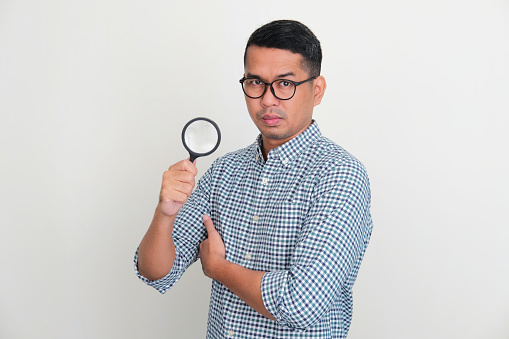 Adult Asian man looking at camera with suspecting expression while holding a magnifying glass