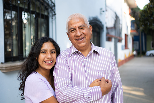 Portrait of grandfather and granddaughter smiling while standing in the street. The granddaughter is hugging the grandfather. They are looking at the camera. High quality photo