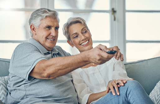 Senior couple, smile and hug watching TV for entertainment on living room sofa with remote at home. Happy elderly man and woman relaxing on couch streaming movies, shows or media at the house