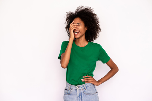 Portrait of happy young woman laughing covering face with hand over white background. African American lady wearing green T-shirt and jeans having fun. Fun and humor concept