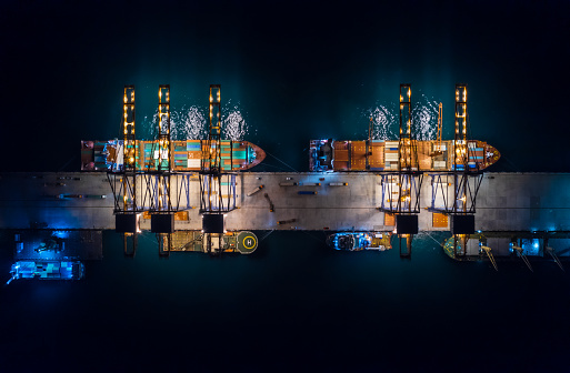 Aerial top view of international port with crane loading containers in import export business logistics. Container loading and unloading cargo freight ship in deep sea at night.