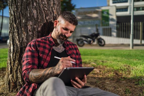 istock Tattooed man sitting on the grass, leaning on tree trunk and happily working on his tablet 1447587014