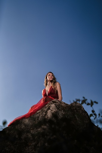 A vertical shot of an attractive female in a long red dress sitting on a rock