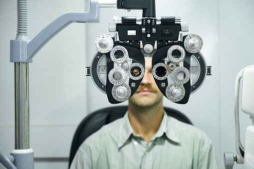 Smart handsome man having eye test, visual examination, check by a professional optician using clinical testing machine phoropter for making new eyeglasses at optic store.