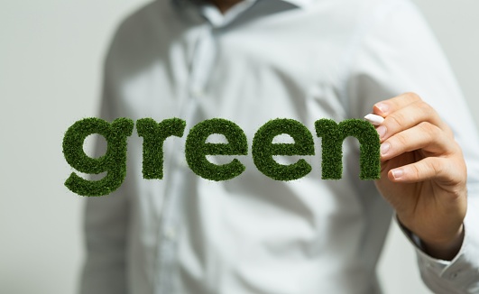 A Caucasian businessman formally dressed pointing with a pen toward a 3D illustration 'green' in grass