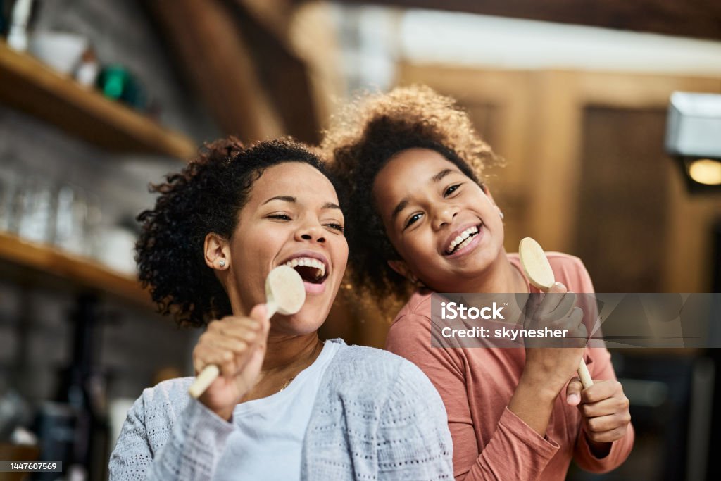 Happy black mother and daughter singing with wooden spoons at home. Happy African American mother and her small girl having fun while singing on wooden spoons in the kitchen. Singing Stock Photo