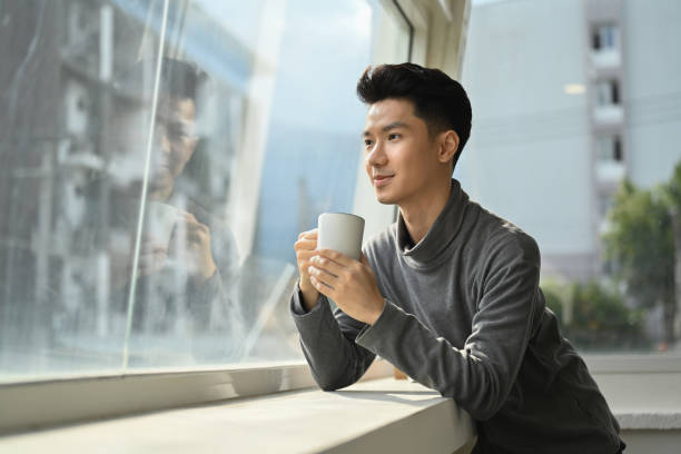 istock Pleasant asian man enjoying his morning coffee while standing near office window overlooking cityscape. 1447609736